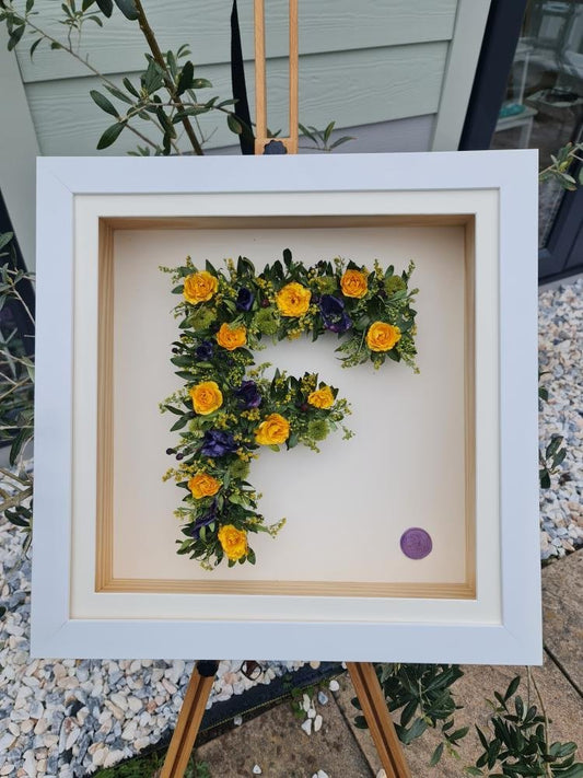 Framed funeraln wreath letter 'F' made of flowers and greenery on a pebbled surface.