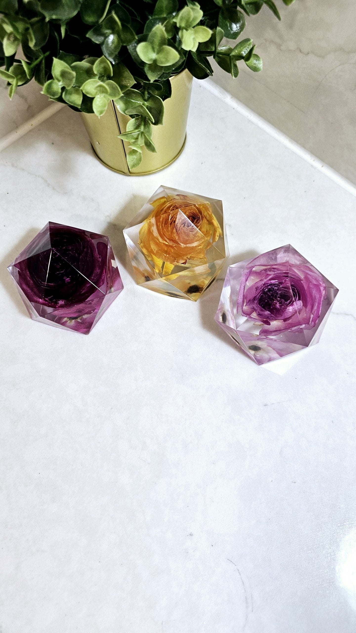 Three colourful resin crystal-like paperweights full of flowers on a white surface with a plant in the background.