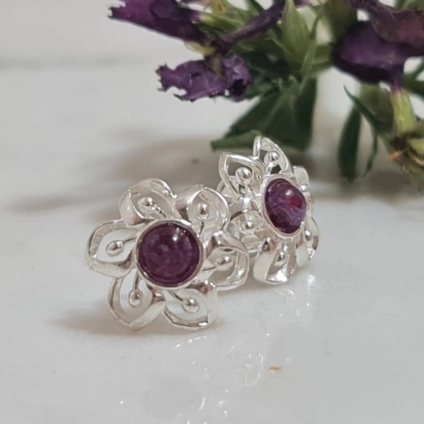 Silver earrings with purple flower preserved on a white surface with purple flowers in the background