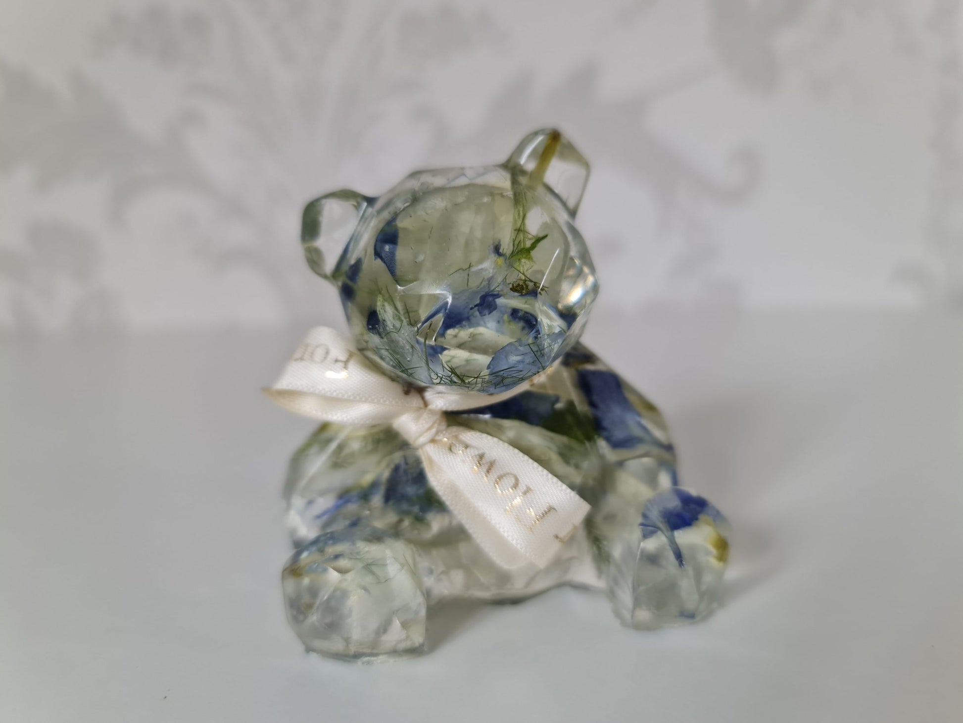 resin figurine of a bear with blue and white petals preserved within it and white ribbon on a light gray background
