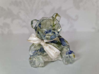 resin figurine of a bear with blue and white petals preserved within it and white ribbon on a light gray background