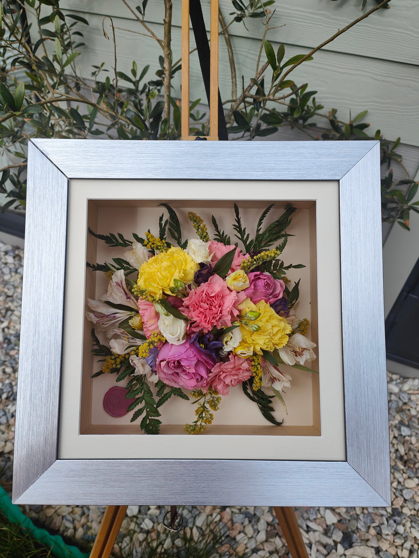 Framed bouquet of colorful pressed flowers on a stand with greenery in the background