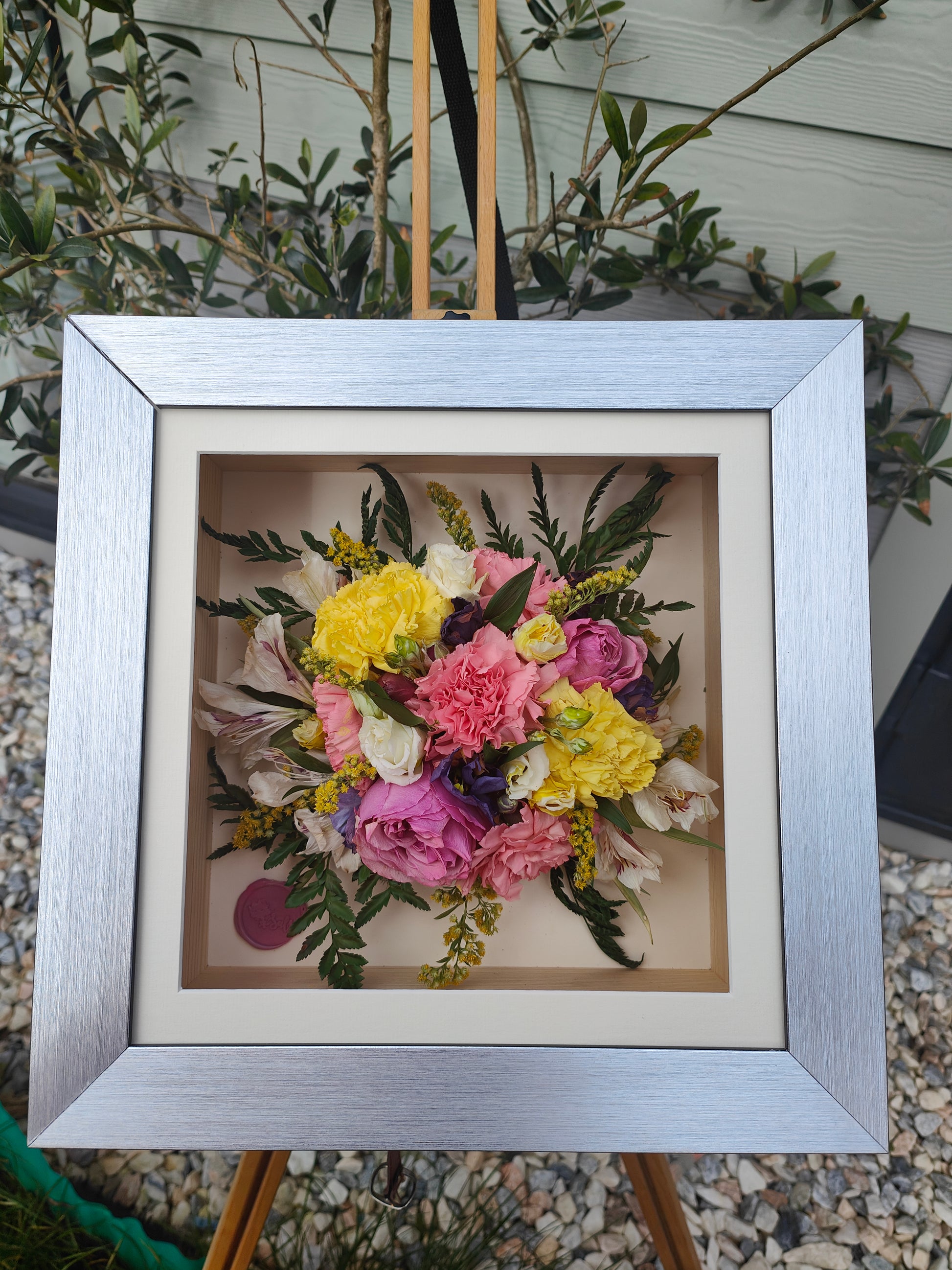 Framed bouquet of colorful pressed flowers on a stand with greenery in the background