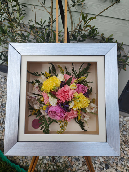 Framed bouquet of colorful pressed flowers on a stand with greenery in the background