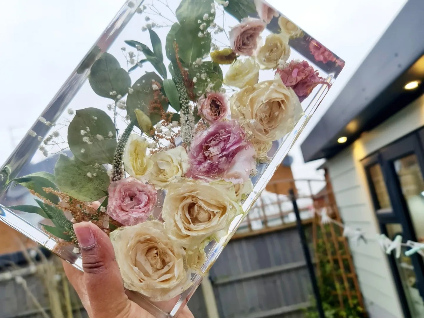 Clear transparent resin rectangle  block with pressed flowers, held by a hand outdoors.