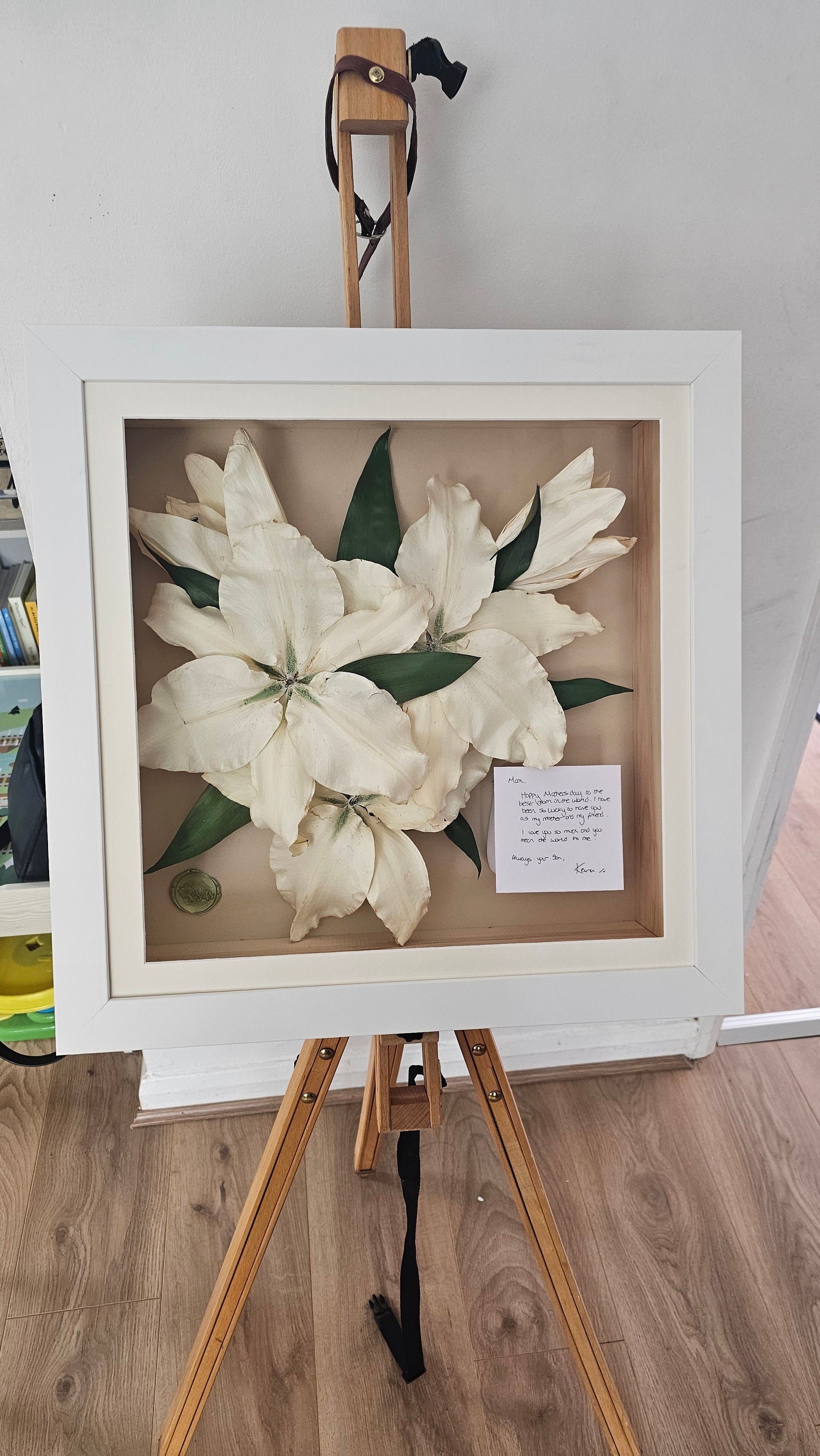 Preserved and Framed funeral lillies on a stand with a wooden floor and white wall background