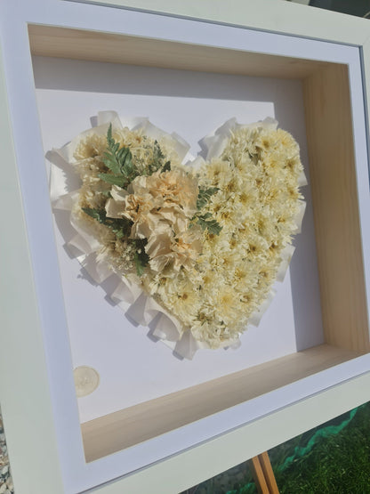 Heart-shaped arrangement of beige memorial flowers from a babies funeral in a white frame.