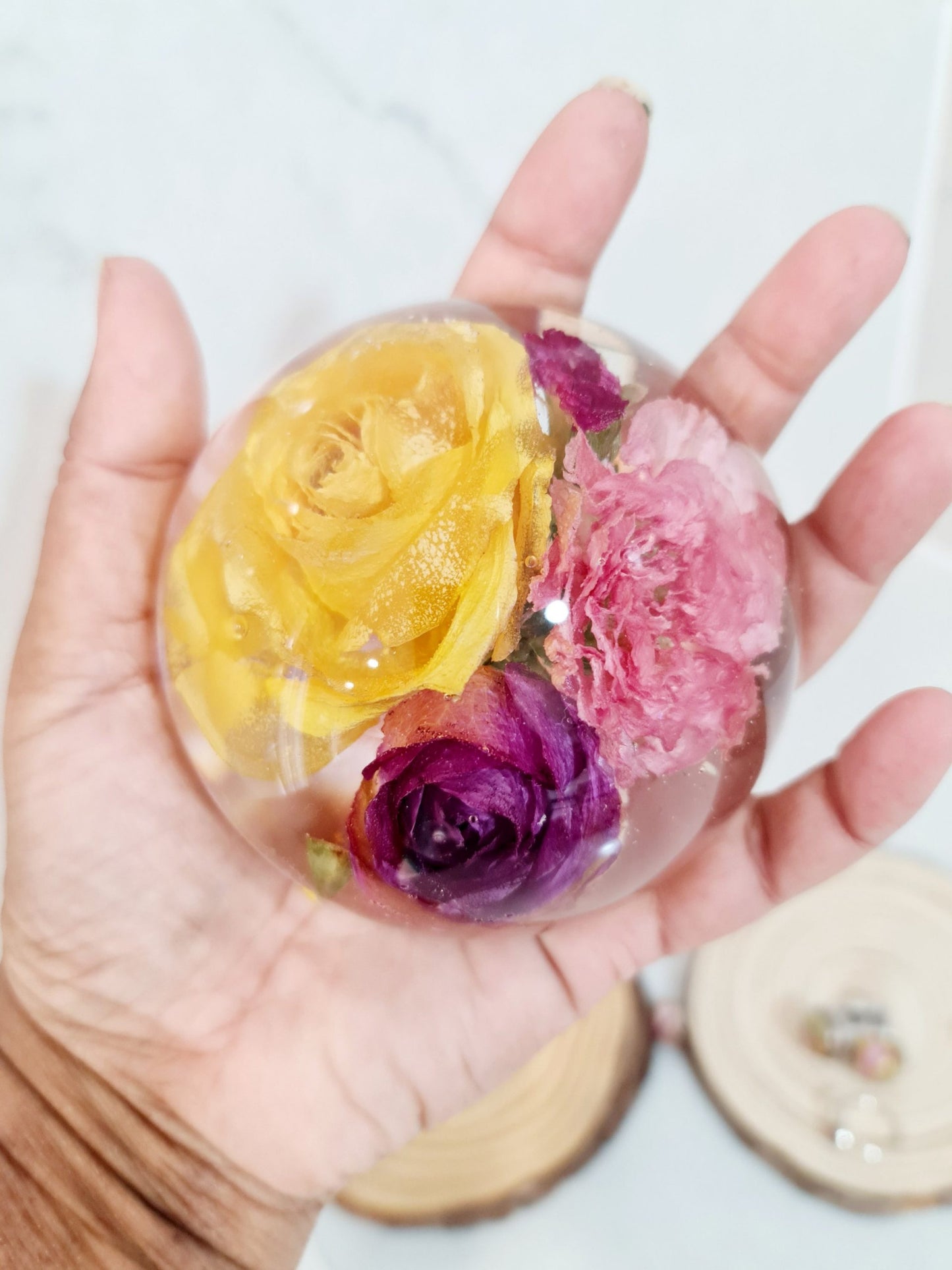 Hand holding a clear resin dome with yellow, pink, and purple flowers on a light background.