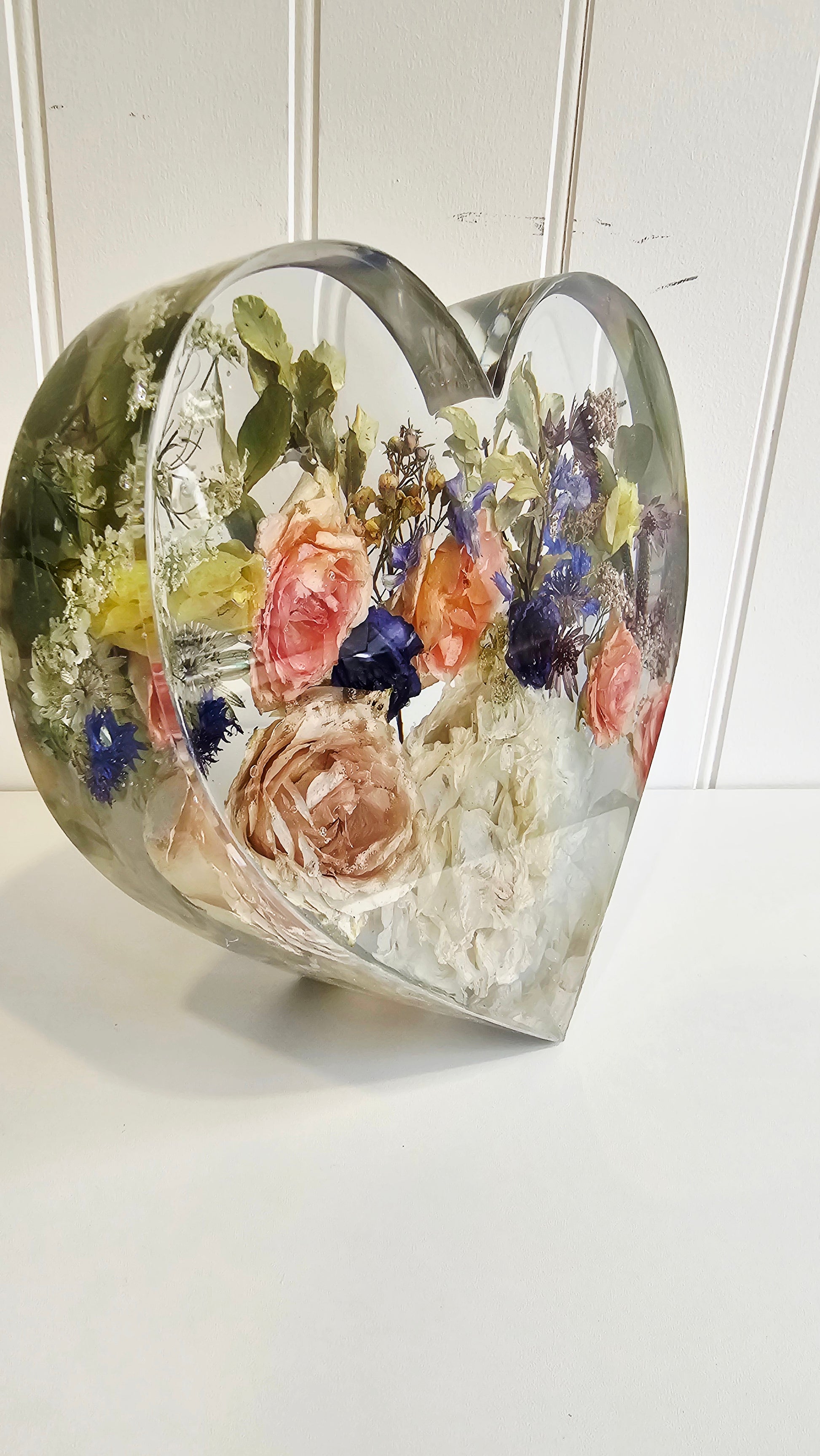 Side view of a standing heart made of clear resin with colourful flowers preserved withing it on a white background, a memory keepsake