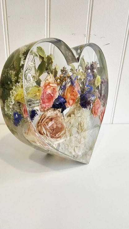 Side view of a standing heart made of clear resin with colourful flowers preserved withing it on a white background, a memory keepsake