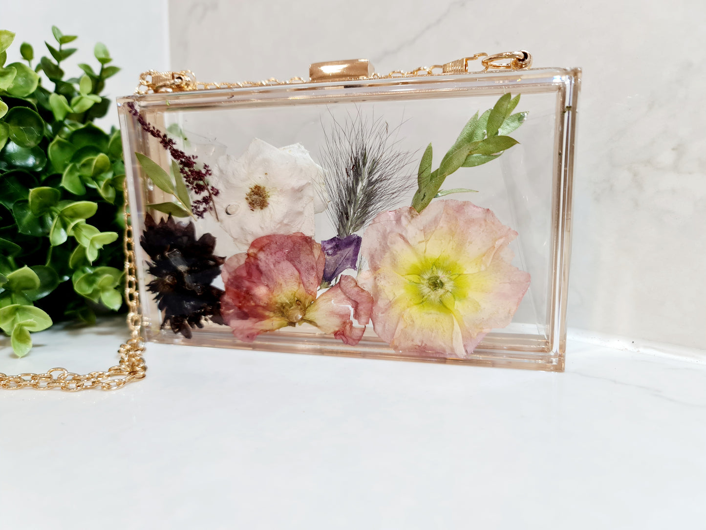 Clear clutch with pressed flowers on a white surface with greenery in the background