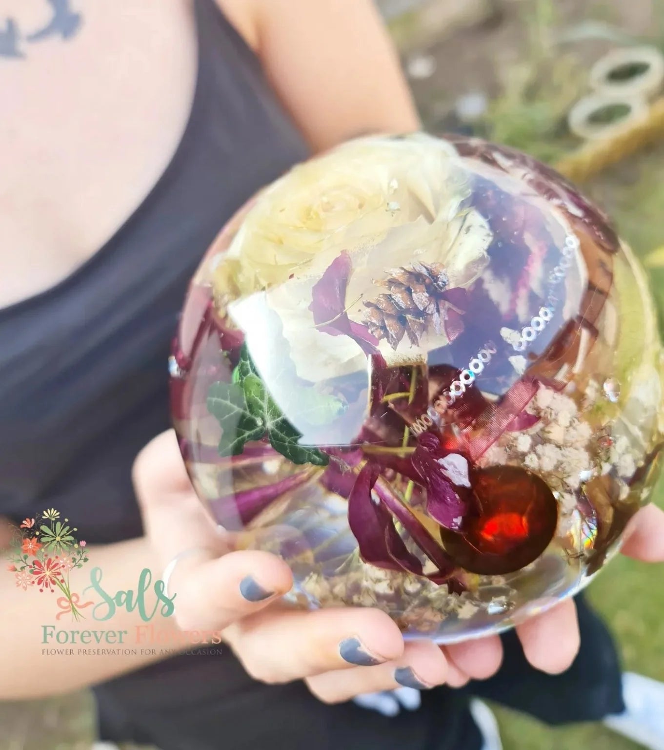 Person holding a transparent resin sphere with embedded wedding flowers and leaves, outdoors. close up view at an angle