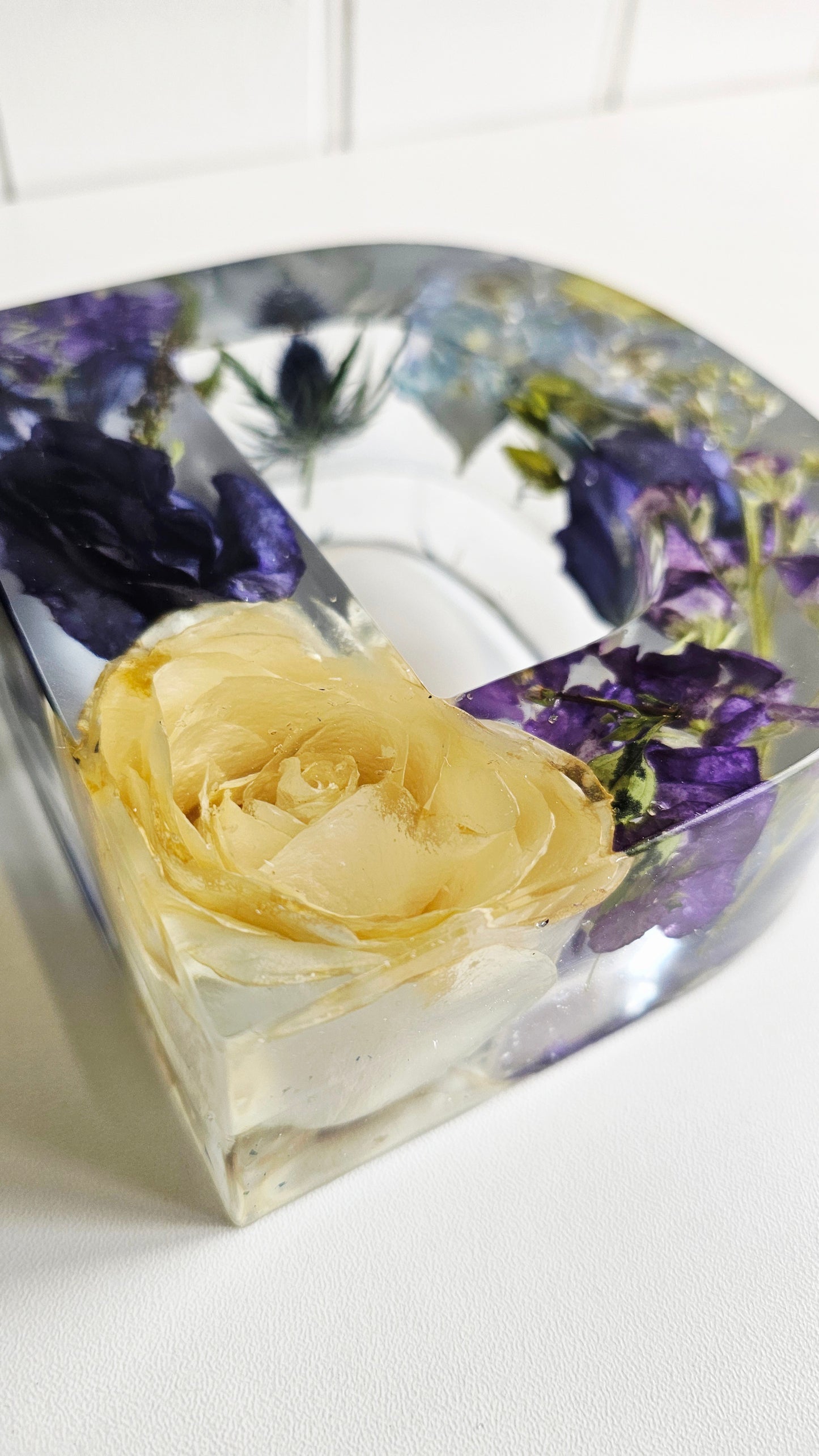 Decorative resin letter d with pressed flowers including a white funeral rose and purple blossoms on a white background. angled view close up