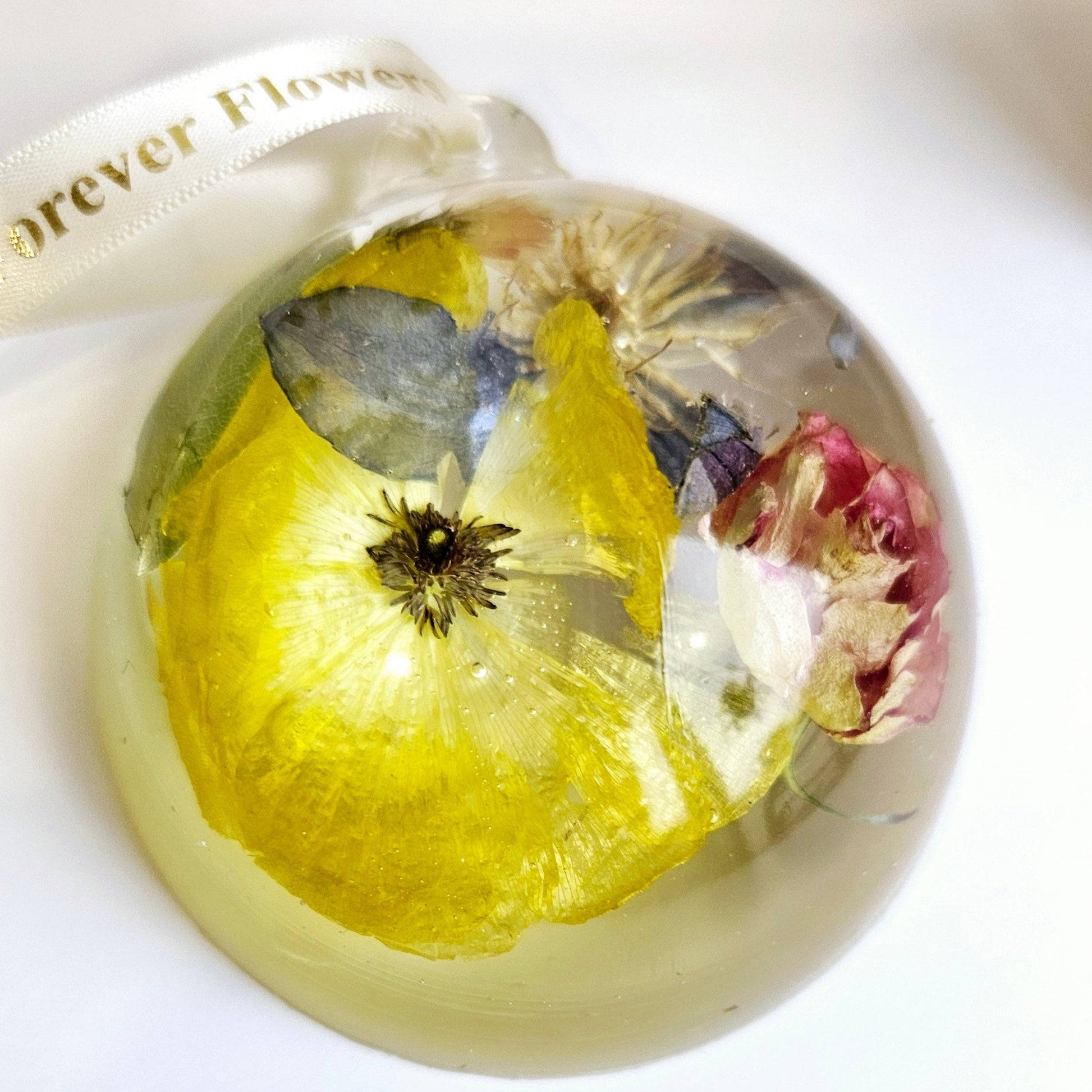 Decorative resin bauble keepsake with yellow liquid and floating flowers on a white background