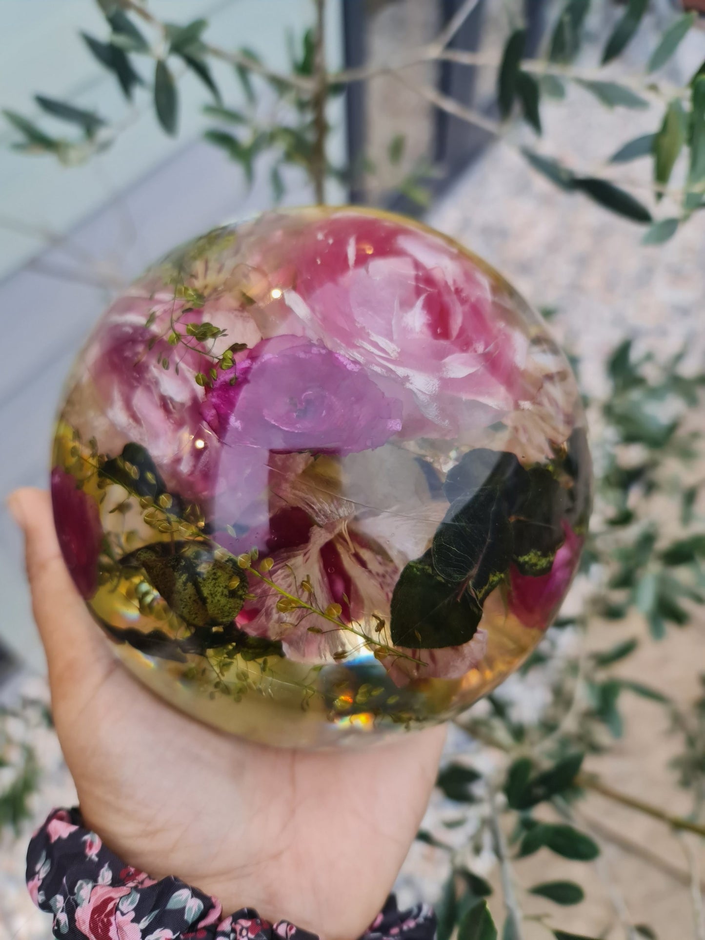 Hand holding a transparent resin sphere with preserved floral and leaf bouquet  against a blurred natural background close view