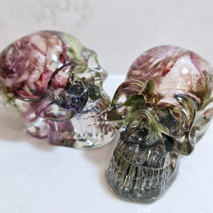 Two decorative resin skulls with floral and greenery inside on a white background