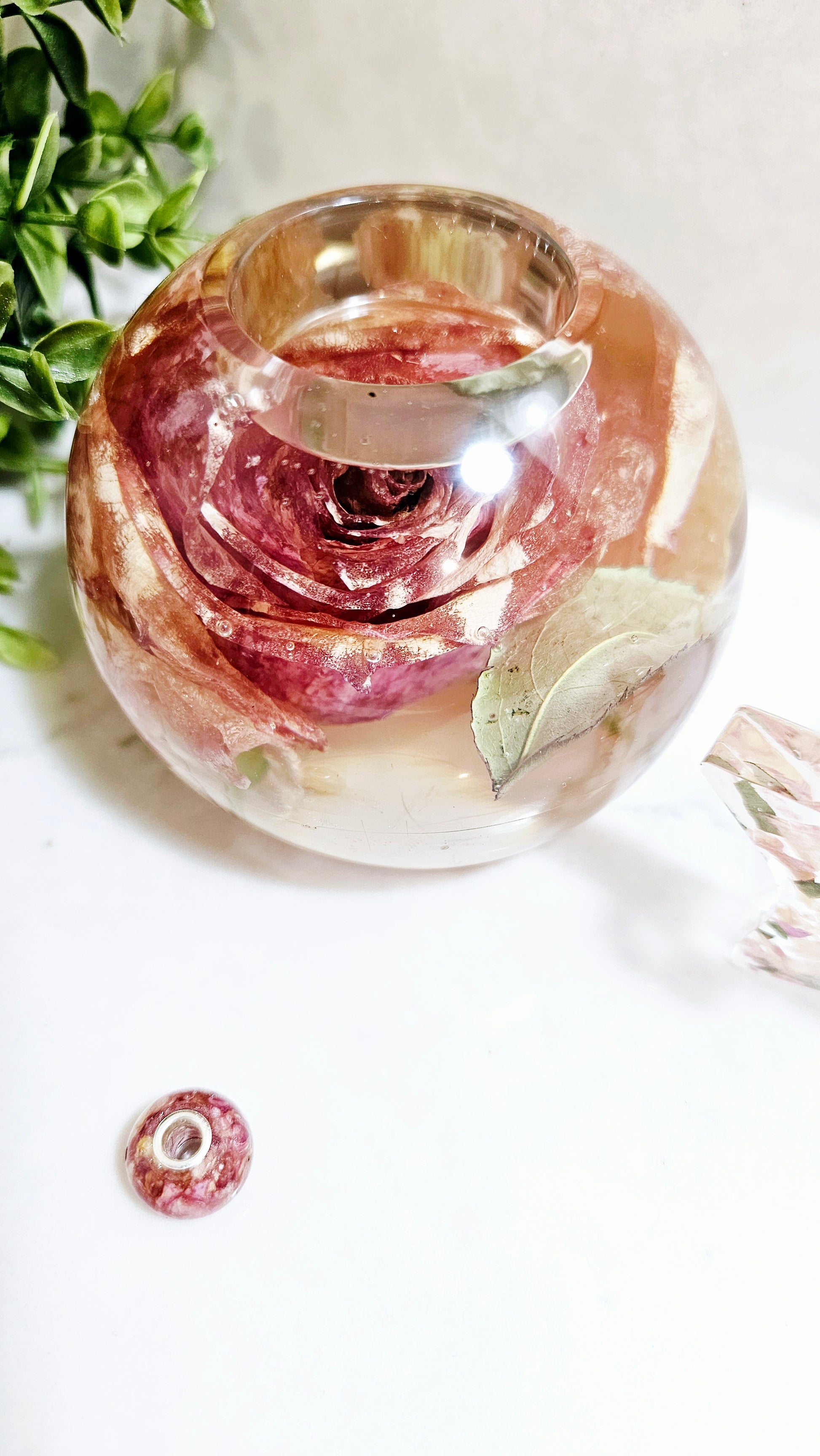 resin sphere tealight holder with a preserved pink rose and leaf on a white surface with greenery.