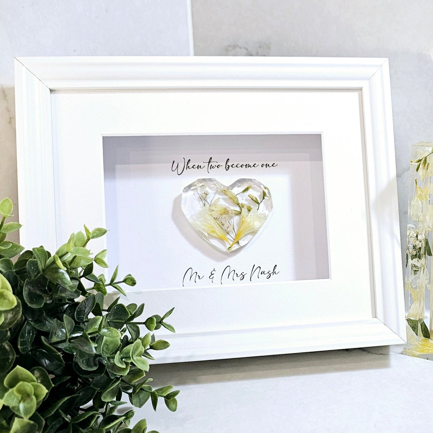Framed artwork with a heart design and text, placed on a surface with a plant and another framed item.